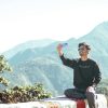A young man captures a selfie with scenic mountains in the background on a sunny day.