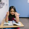 A confident young girl in a classroom giving a thumbs-up while reading a book.